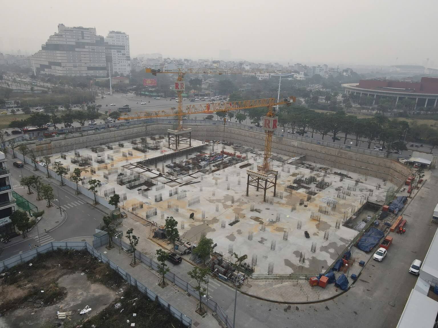 Tiến độ thi công Vinhomes Nguyễn Trãi - Cập nhật mới nhất 11 Hình ảnh từ trên cao của công trường Vinhomes Nguyễn Trãi trong giai đoạn thi công móng, với nền bê tông và vật liệu xây dựng, cùng khung cảnh thành phố Hà Nội mờ sương xung quanh.